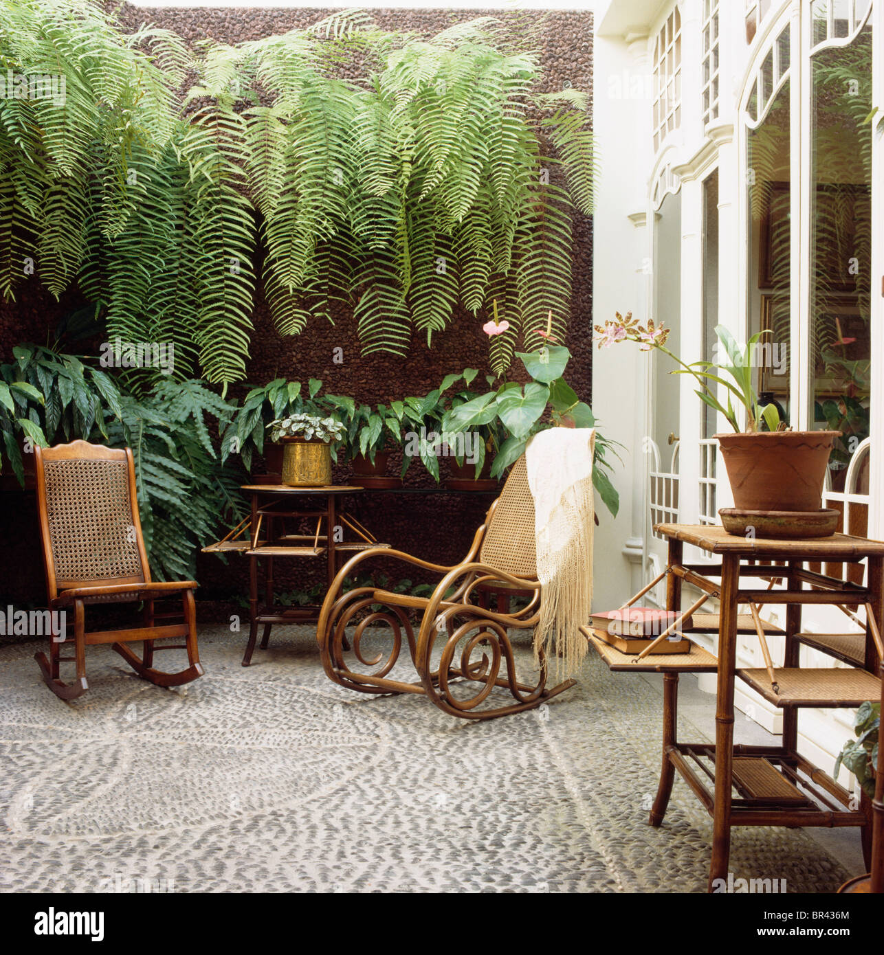 Antique Bentwood a dondolo sedie sotto la parete con lussureggianti felci nel cortile con il mosaico di ciottoli di pavimentazione e tabella di bambù Foto Stock