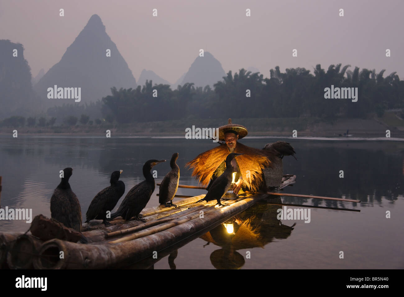 Fisherman indossando la paglia umettare la pesca con i cormorani su zattera di bambù sul Fiume Li al crepuscolo, Yangshuo, Guangxi, Cina Foto Stock