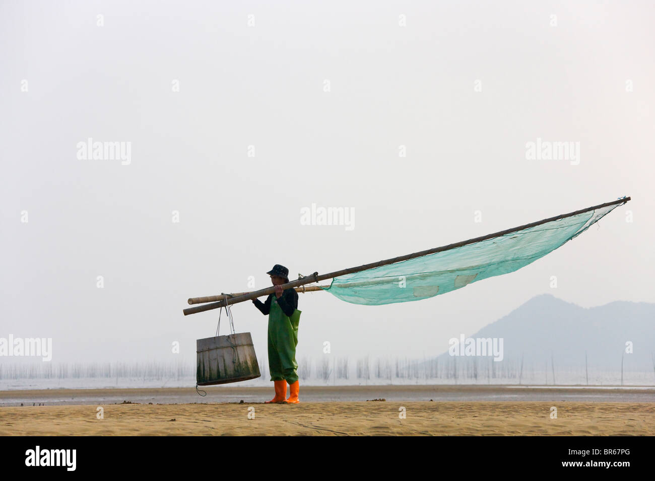Fisherman trasporto netto di pesce sulla spiaggia, canne di bambù per essiccare le alghe in distanza, ad est del Mar della Cina costa, Xiapu, Fujian, Foto Stock