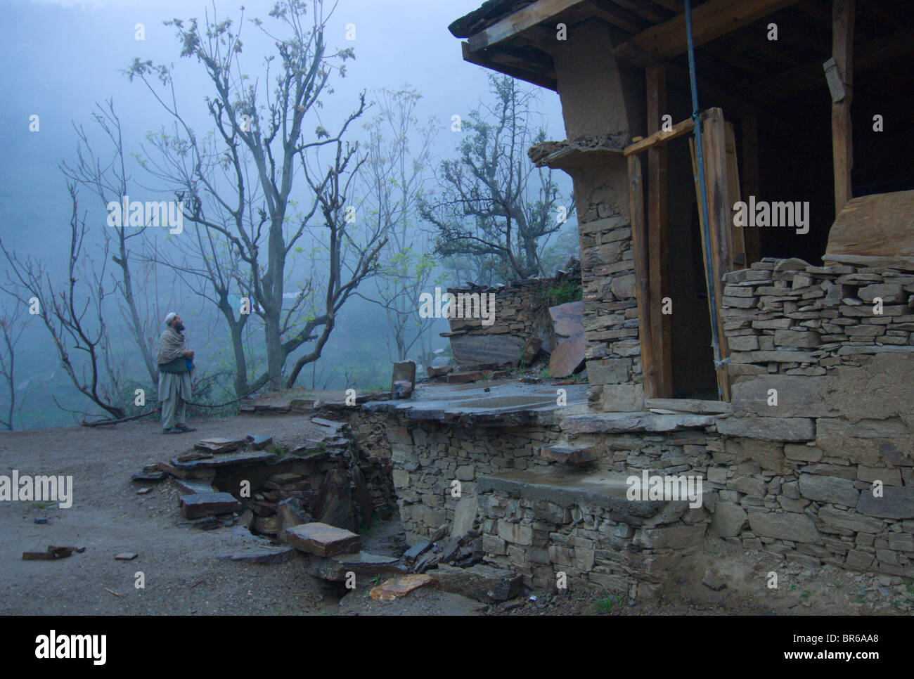 Un Pashtun l uomo sta sotto la pioggia e nebbia al di fuori di una casa danneggiata nel terremoto-lui e la sua famiglia hanno appena arrivato indietro Foto Stock