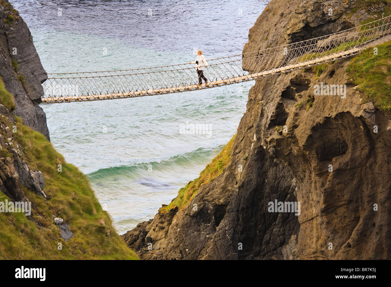 Giovane donna che attraversa il Carrick-a-Rede ponte di corde nei pressi del villaggio di Ballintoy sulla contea costa di Antrim, Irlanda del Nord Foto Stock