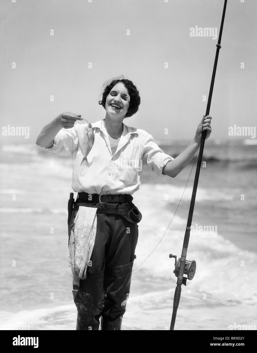 1920s 1930 Donna sorridente IN PIEDI IN OCEAN SURF INDOSSANDO WADERS GOMMA TENENDO IL PESCE E FLY CANNA DA PESCA Foto Stock