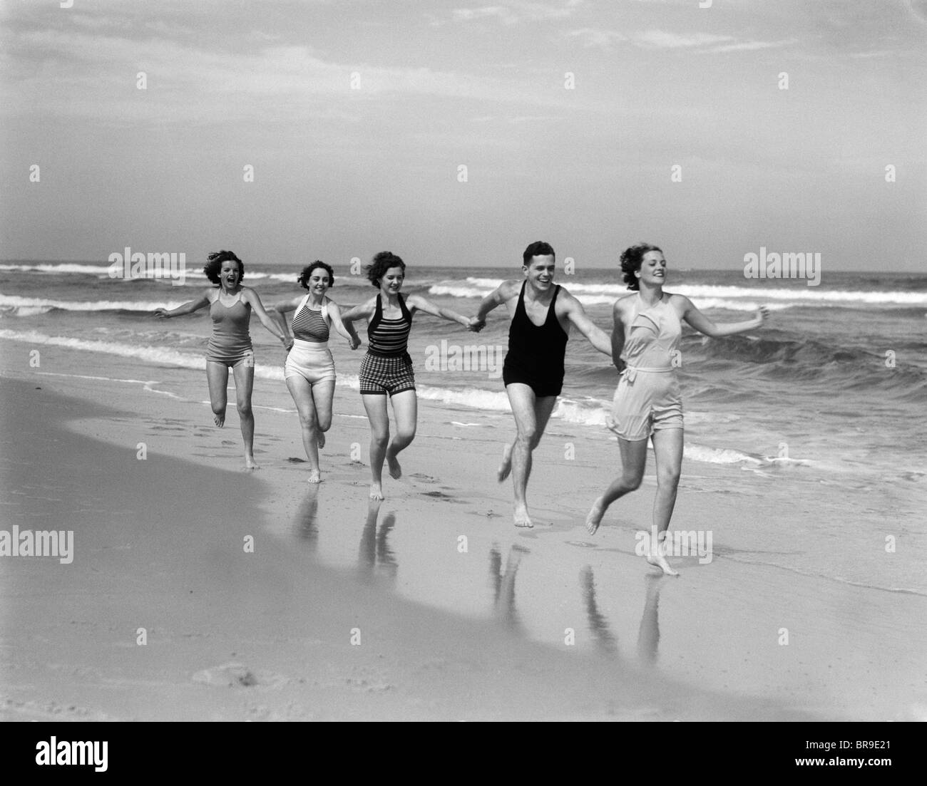 1930 Quattro donne e un uomo che corre sulla spiaggia tenendo le mani Foto Stock