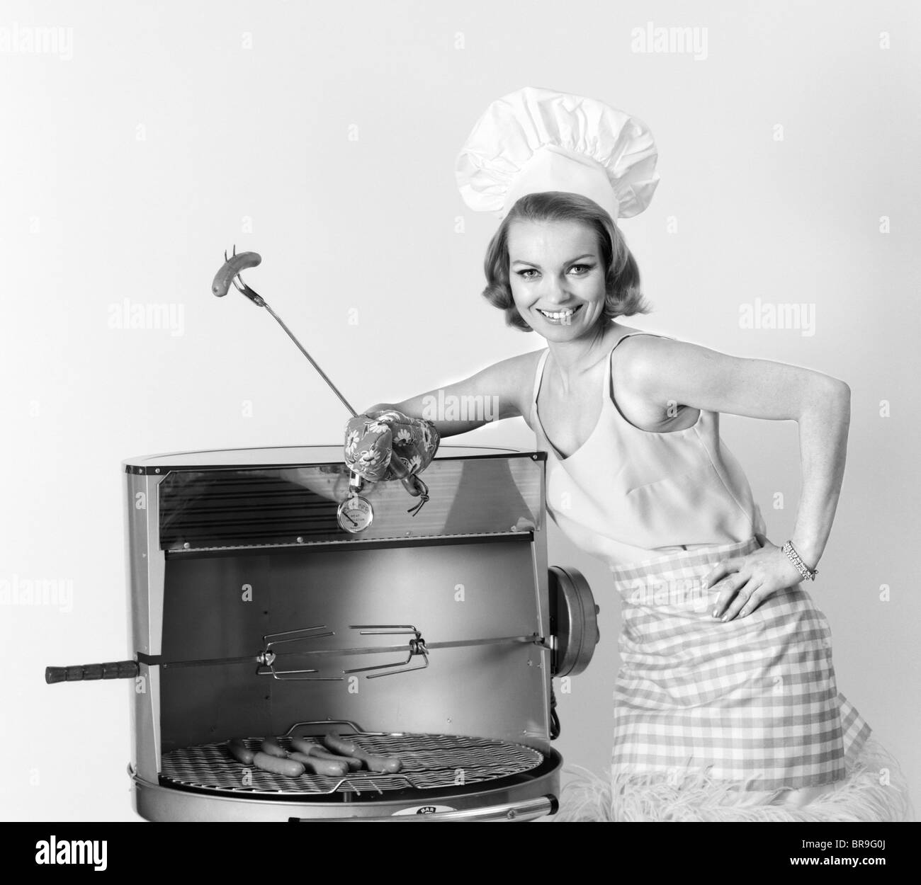 Anni sessanta donna che indossa lo chef HAT permanente al barbecue azienda una forcella con HOT DOG Foto Stock