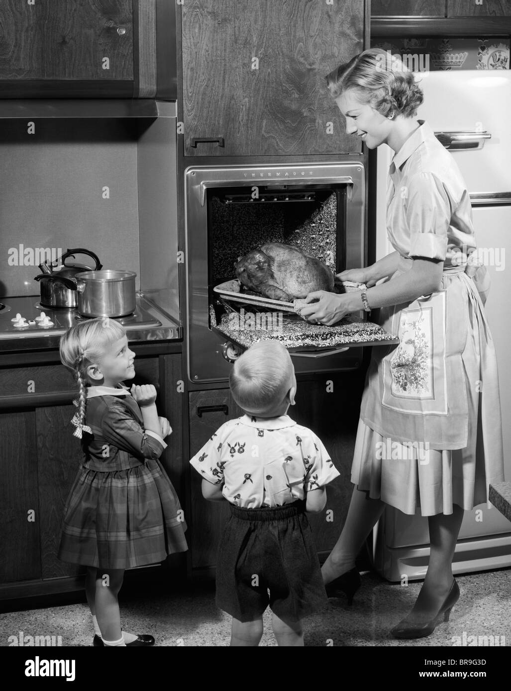 Anni Cinquanta la figlia e il figlio guardando la madre staccare arrosto di tacchino del Ringraziamento a cena da cucina elettrica forno Foto Stock
