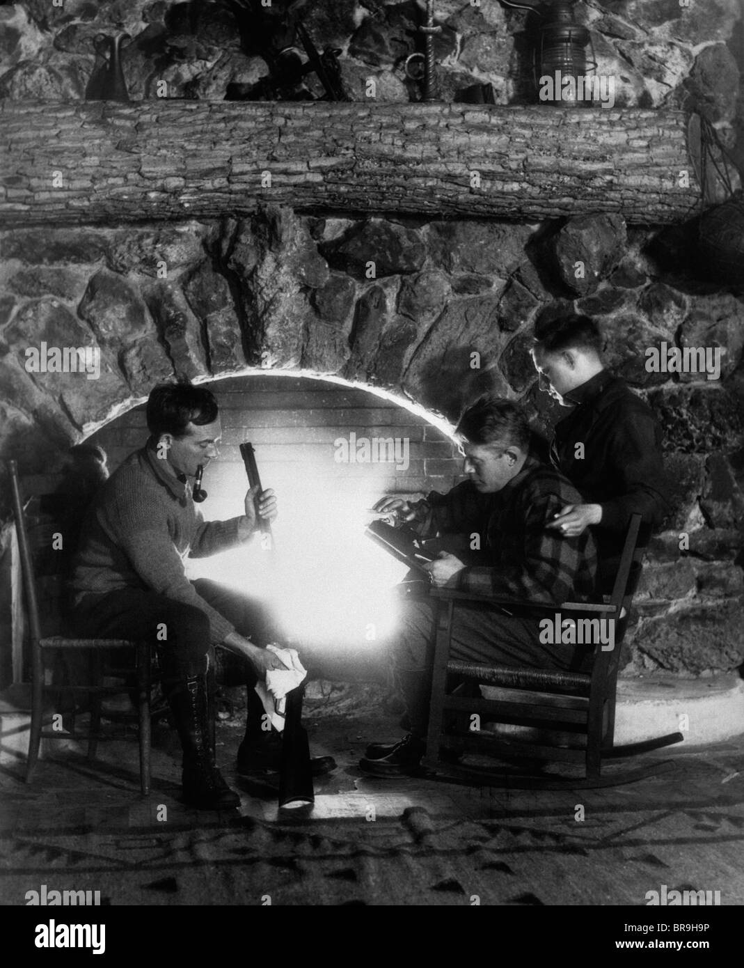 1920s 1930 tre cacciatori di uomini seduti davanti al caminetto in pietra pulizia fucile lucidatura di caricamento di polvere da sparo nel fucile Foto Stock