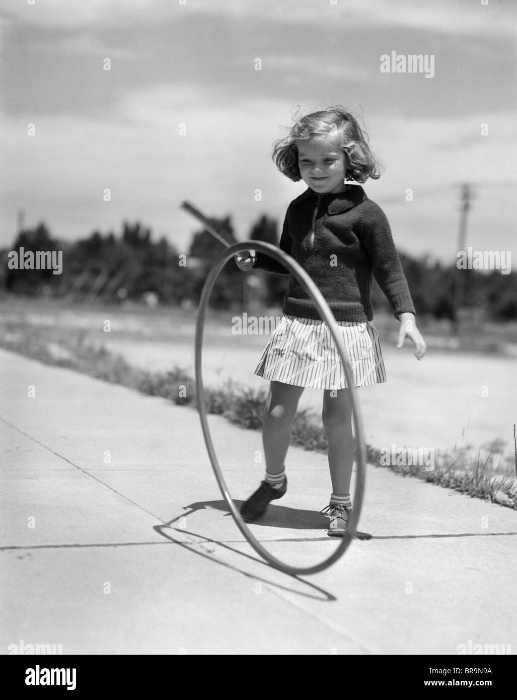 1930s ragazza che gioca con il cerchio e stick sul marciapiede Foto Stock
