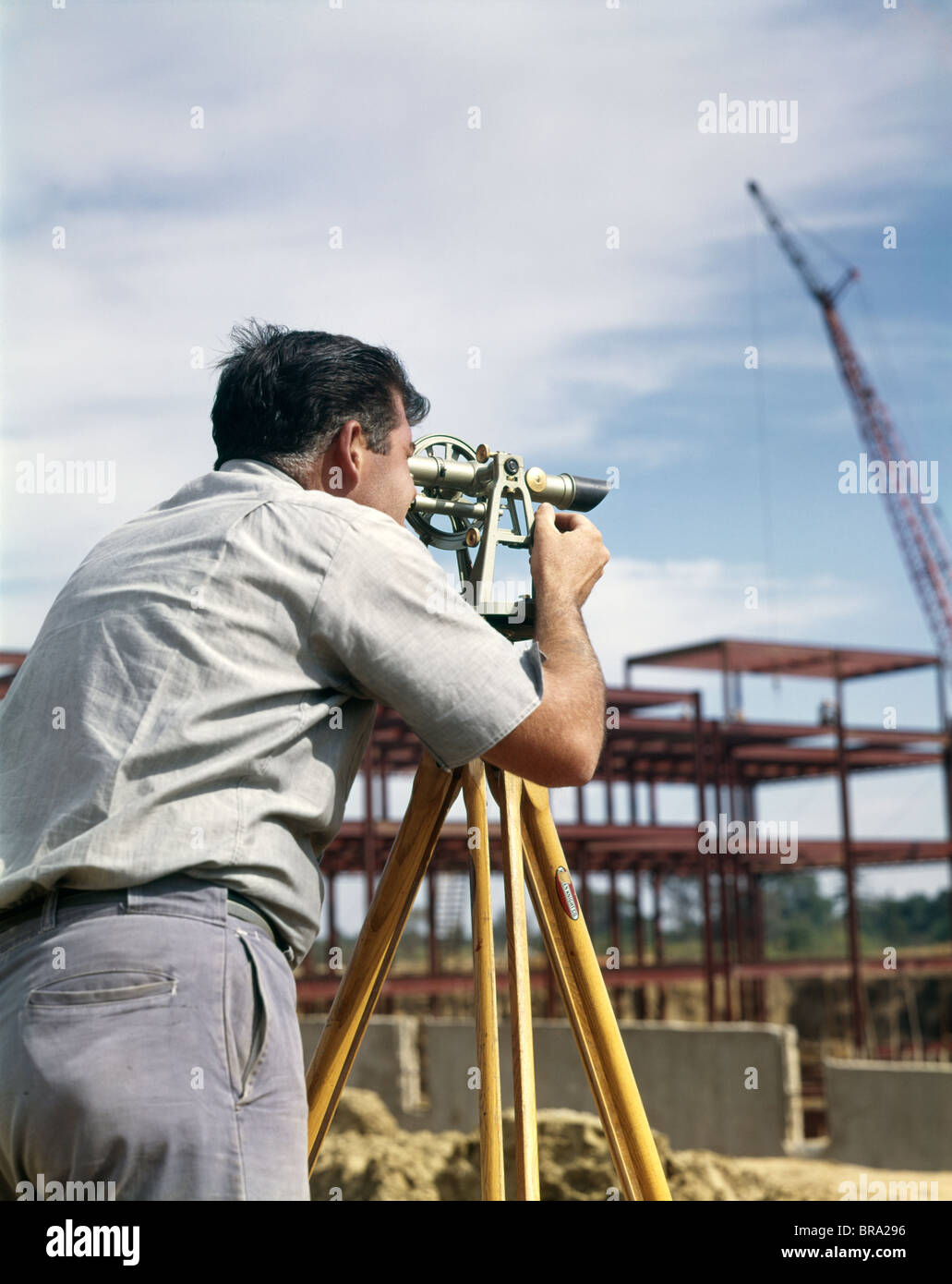 Anni sessanta uomo utilizzando apparecchiature di rilevazione di avvistamento di transito di geometra ingegnere TEODOLITE SITO IN COSTRUZIONE retrò Foto Stock