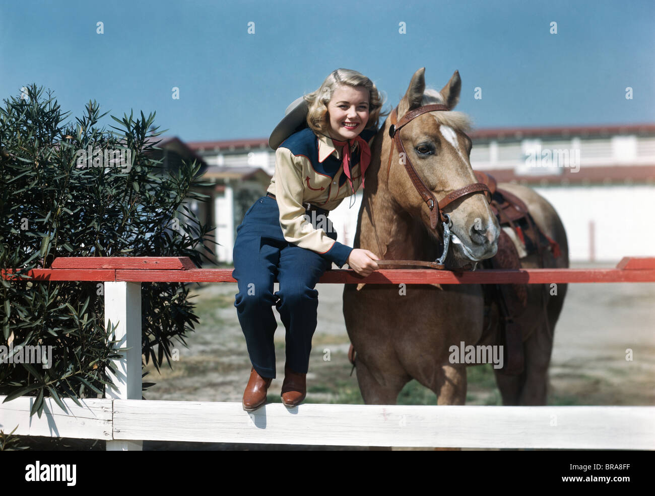 Negli anni quaranta anni cinquanta sorridente ragazza TEEN INDOSSANDO WESTERN COWGIRL OUTFIT seduto sulla recinzione in posa A CAVALLO Foto Stock