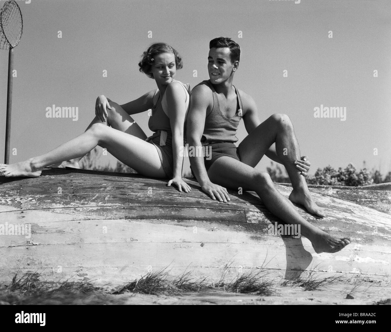 1930s giovane uomo donna sorridente indossando il costume da seduta di schiena su rovesciato la barca di legno Foto Stock