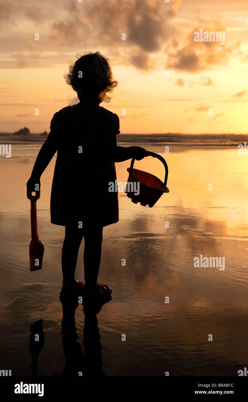 Bambina con benna e vanga stagliano contro il sole di setting su Summerleaze Beach, Bude, UK. Foto Stock