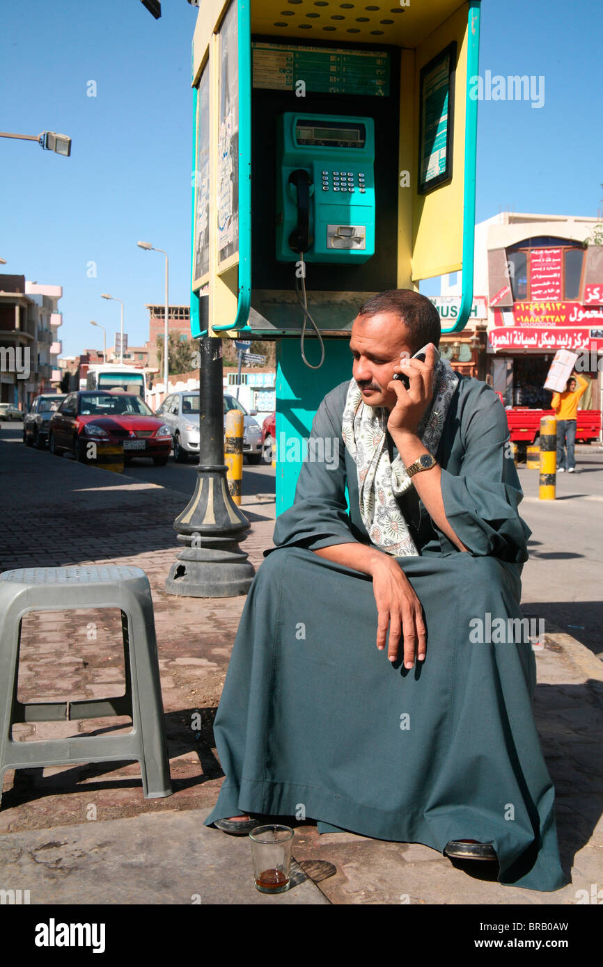 Uomo seduto sotto un telefono pubblico utilizzando un telefono cellulare. Quest'ultimo ha sostituito il primo, Hurghada, Egitto Foto Stock