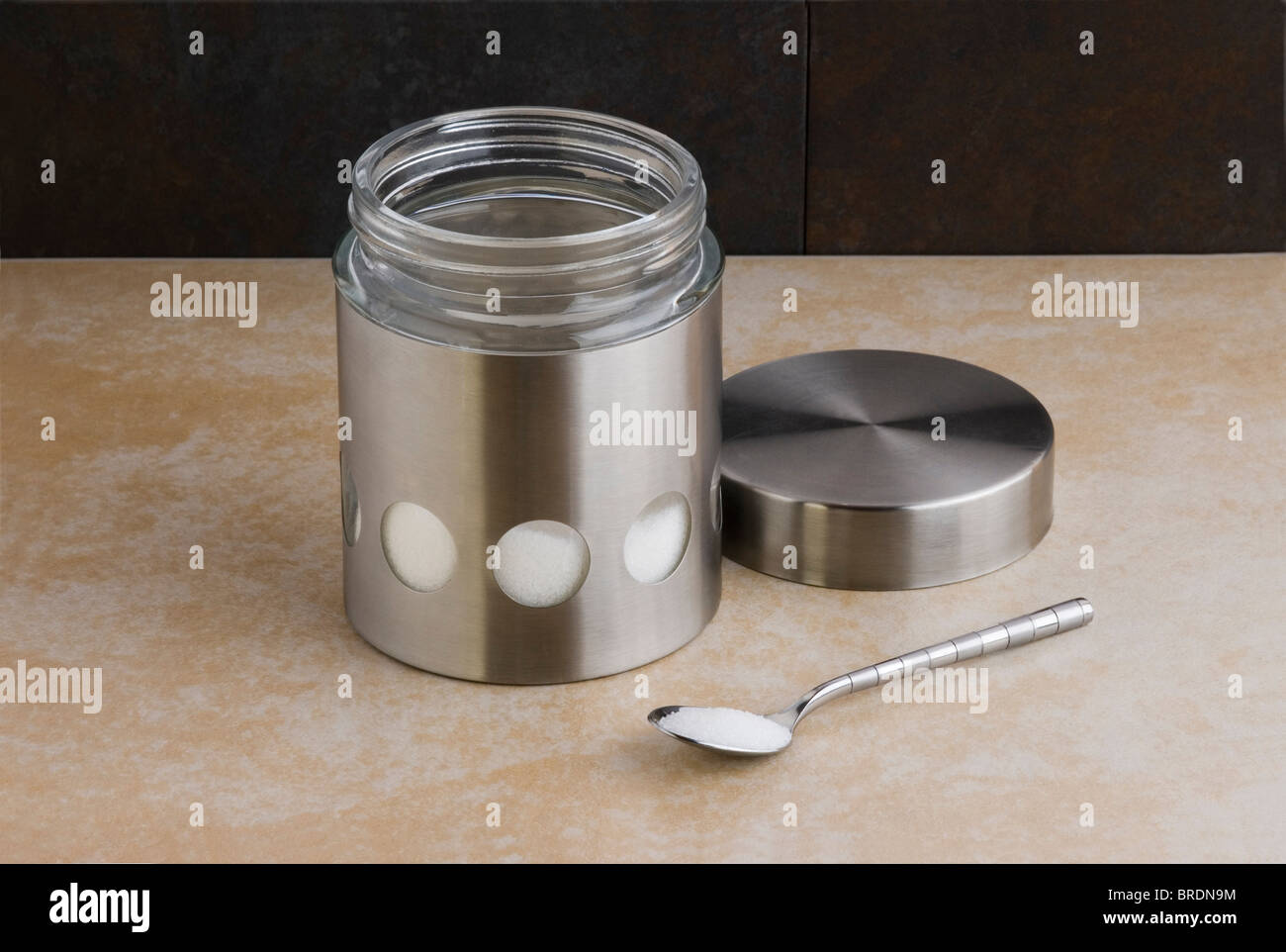 In acciaio inox e vetro barattolo di zucchero, con cucchiaino su un piano di lavoro. Foto Stock