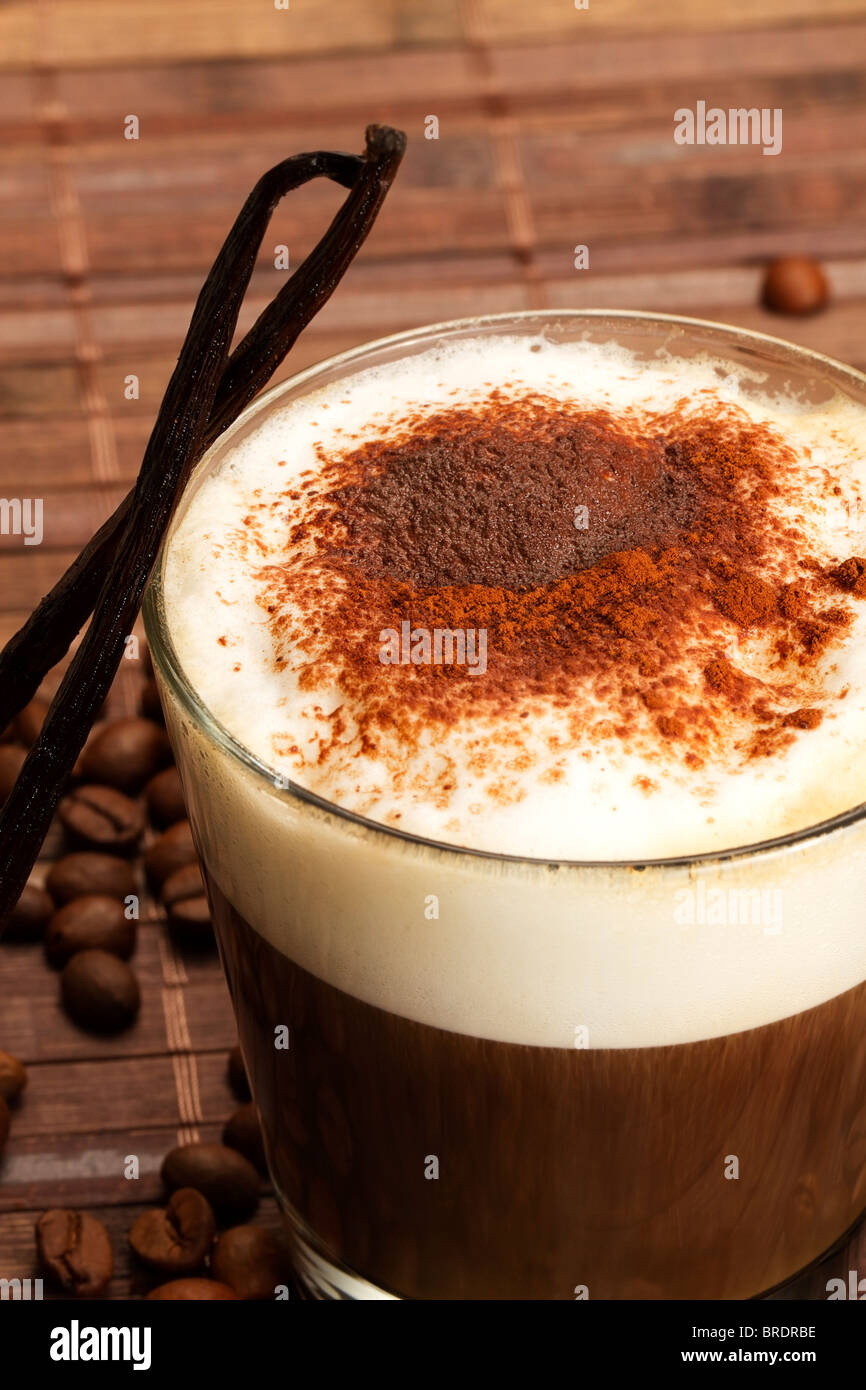 Il caffè con il latte con la schiuma, polvere di cacao e permanente di baccelli di vaniglia su sfondo di legno con i chicchi di caffè Foto Stock