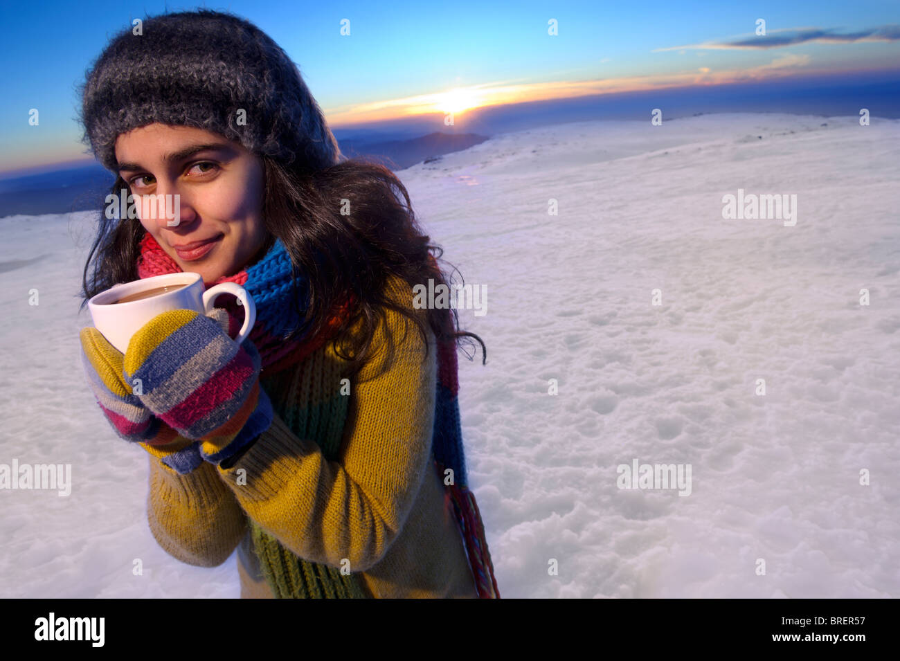 Un sorridente giovane donna detiene una tazza di cioccolato caldo nella neve davanti ad un Cielo di tramonto Foto Stock