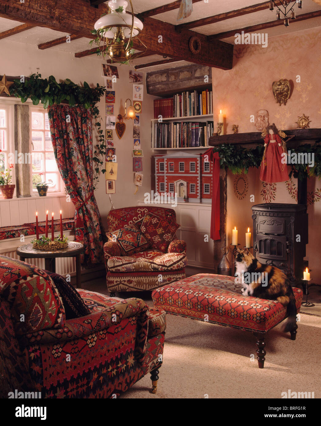 Gatto seduto sul rosso Sgabello imbottito in cottage soggiorno decorato per il Natale con fogliame e candele accese Foto Stock
