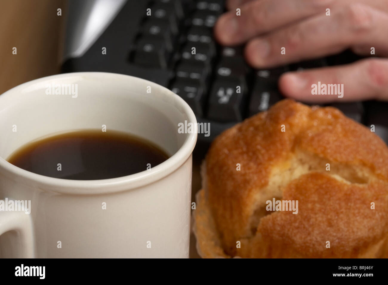 Tazza di caffè nero e un muffin di fronte a una mano mans digitando su una tastiera di computer Foto Stock