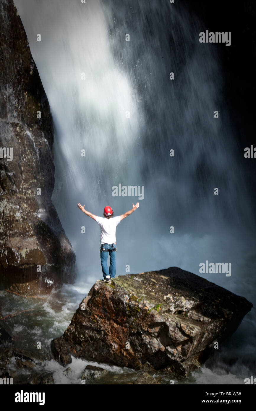 Un giovane uomo tiene le sue braccia aperta permanente alla base di una cascata dopo rappeling down. Foto Stock