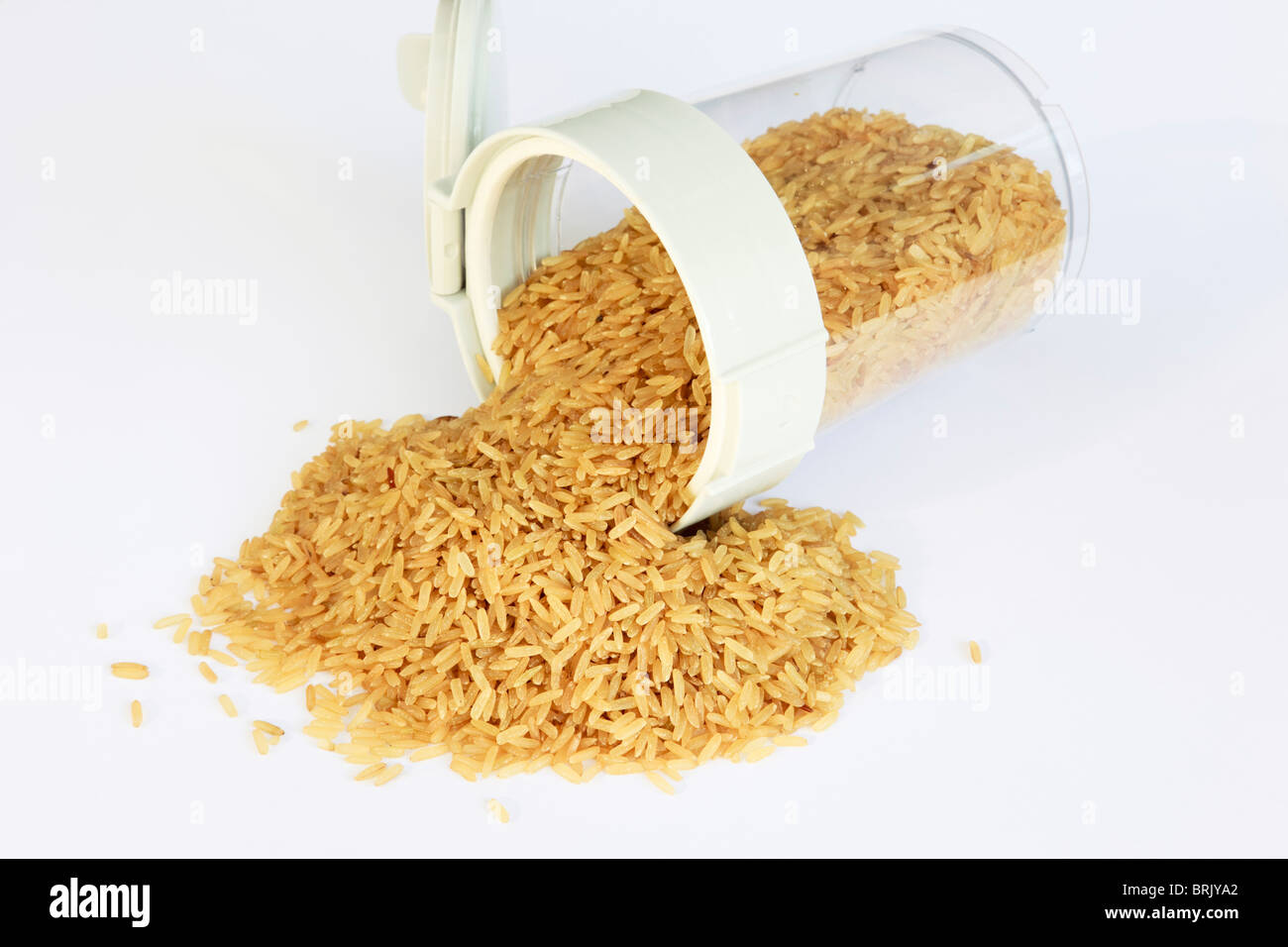 Pila di riso integrale crudo fuoriuscita di un contenitore per alimenti con coperchio aperto su un tavolo bianco Foto Stock