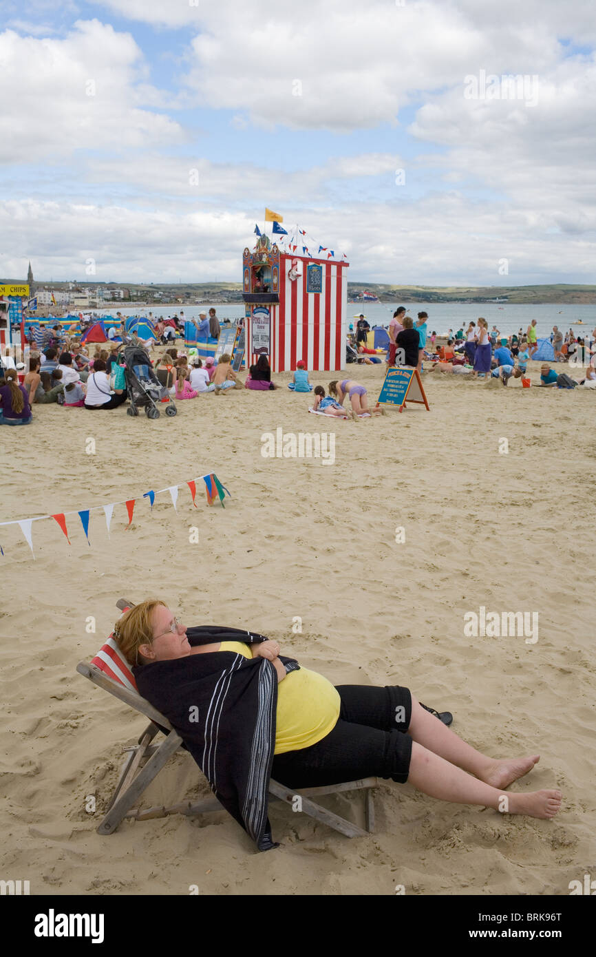 Una grande donna in una sedia a sdraio accanto a Punch & Judy sulla spiaggia di Weymouth. Foto Stock