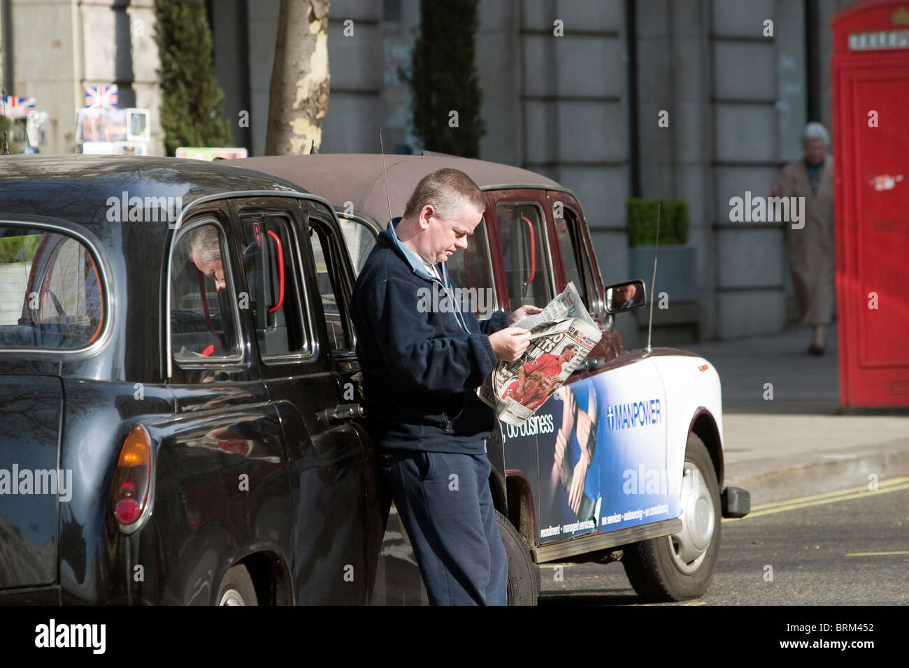 Un tassista leggendo il suo giornale appoggiata contro il suo taxi in London street scene Foto Stock