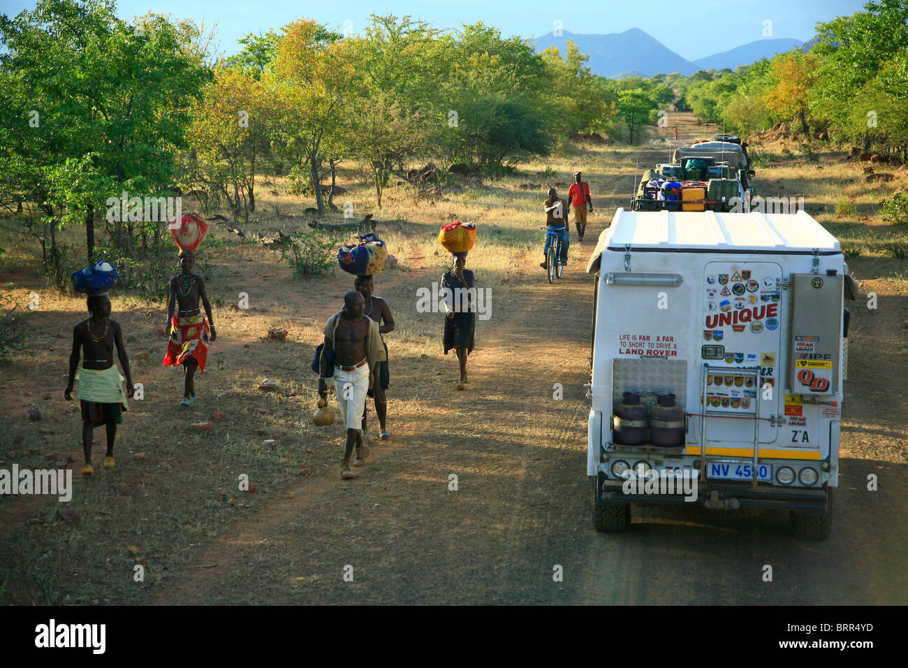 Donne locali a piedi e il convoglio di veicoli 4x4 su una lunga strada diritta in Angola meridionale Foto Stock