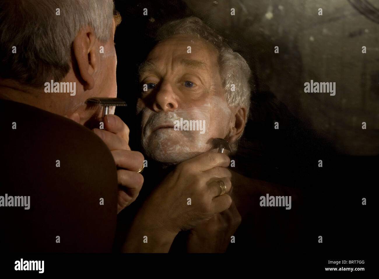 Immagine atmosferica di un pelo grigio l uomo nella sua anni sessanta la rasatura con un vecchio rasoio di sicurezza mentre cercano in un specchio sporco Foto Stock