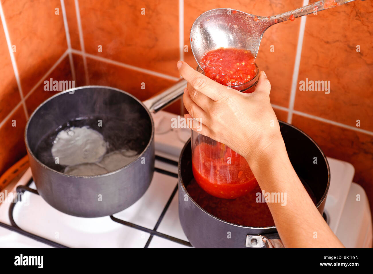 Una donna di mano trattiene un pieno per metà vasetto di vetro mentre riscaldata versare il succo di pomodoro da una siviera, sopra una pentola di ferro. Foto Stock