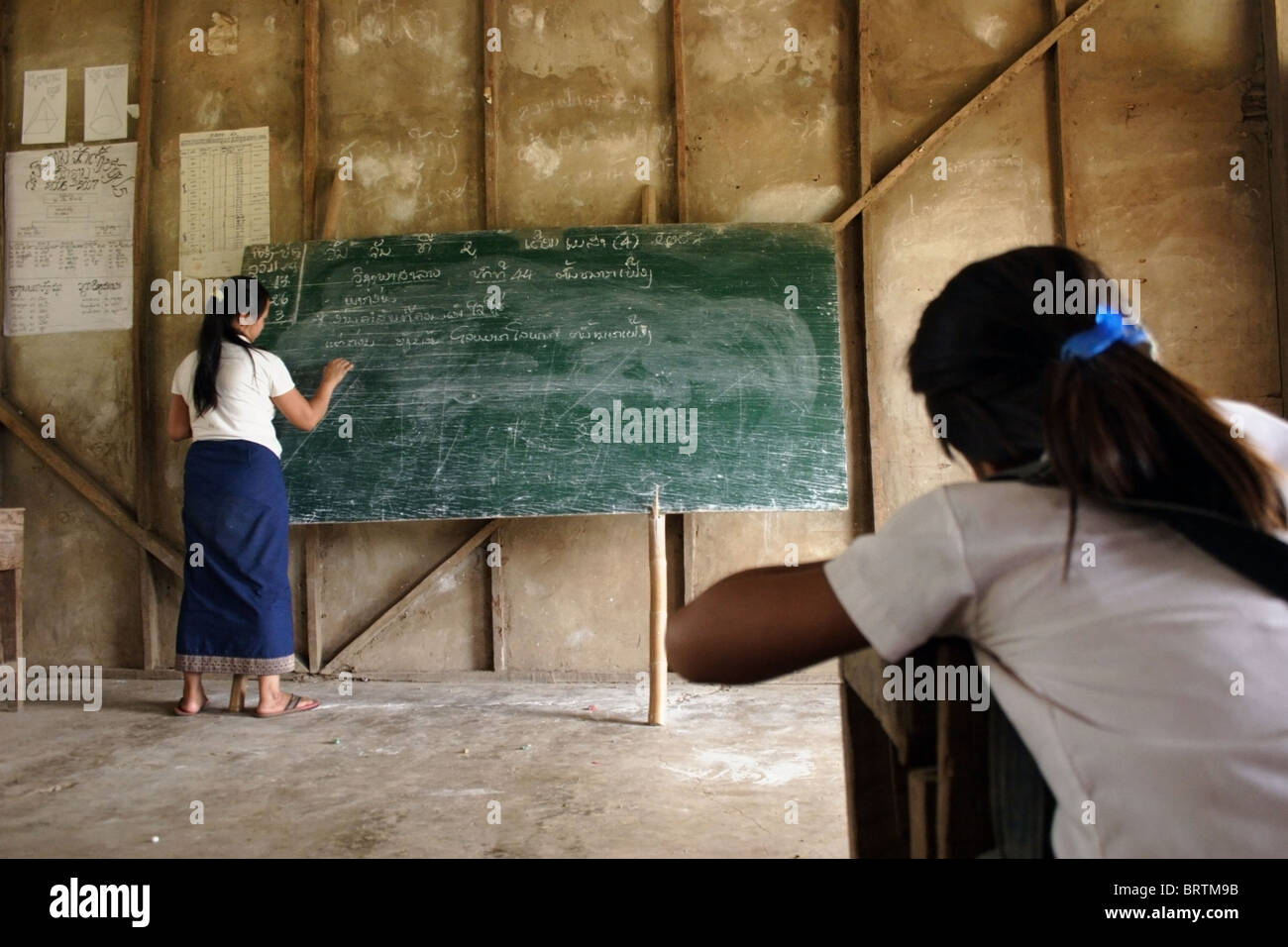 Un insegnante è la scrittura di note su una lavagna verde come una ragazza studente studi presso una scuola primaria di Ban Buamlao, Laos. Foto Stock