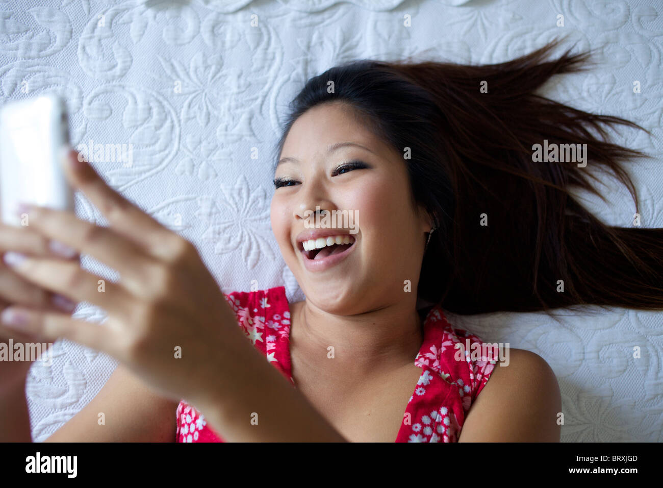 Sorridente razza mista ragazza adolescente posa sul letto e di messaggistica di testo sul telefono cellulare Foto Stock