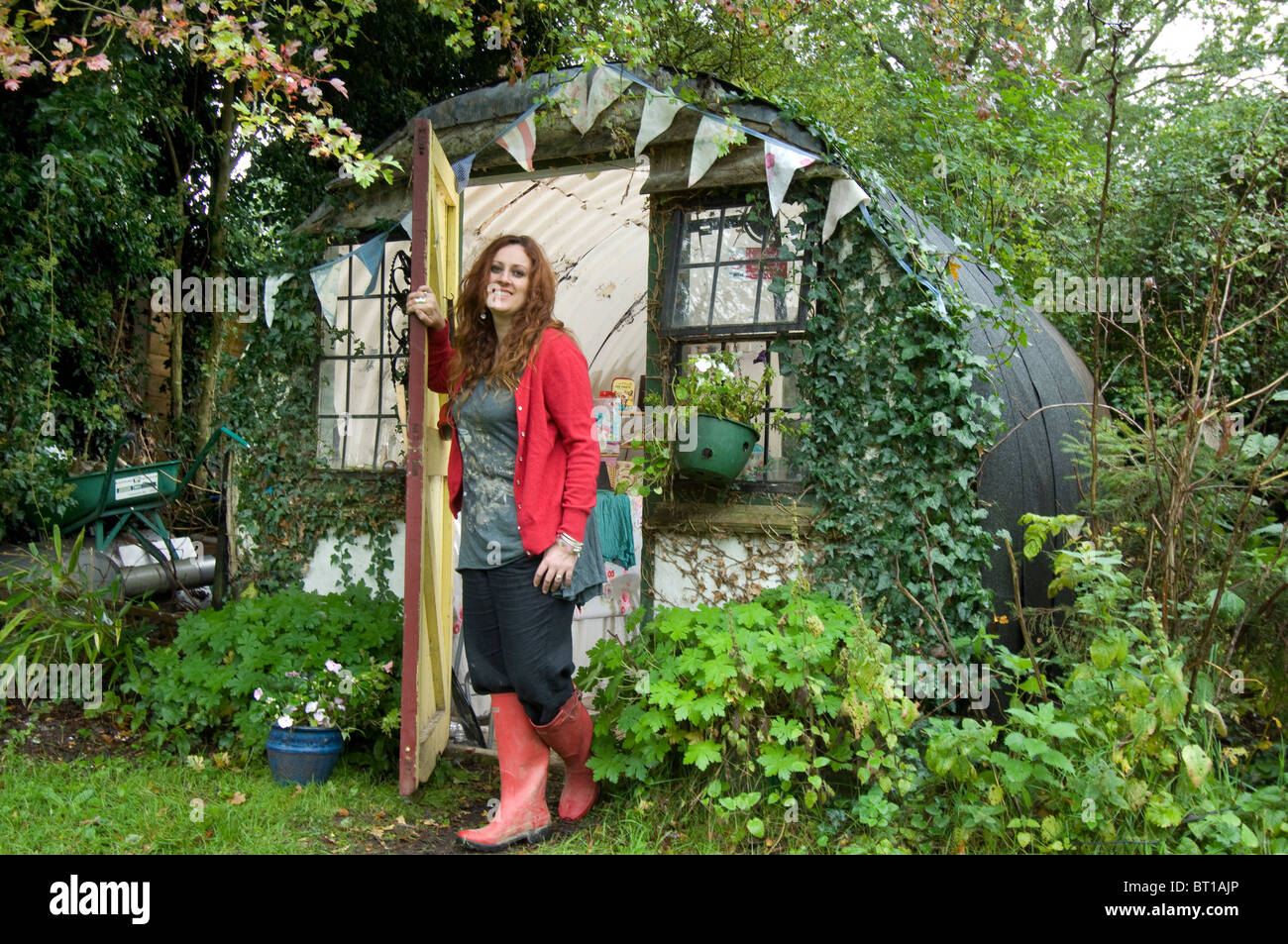 Woman in Red stivali da pioggia alla porta del suo ufficio e della sua Tettoia da giardino realizzato a partire da un vecchio ferro corrugato bomb shelter Foto Stock