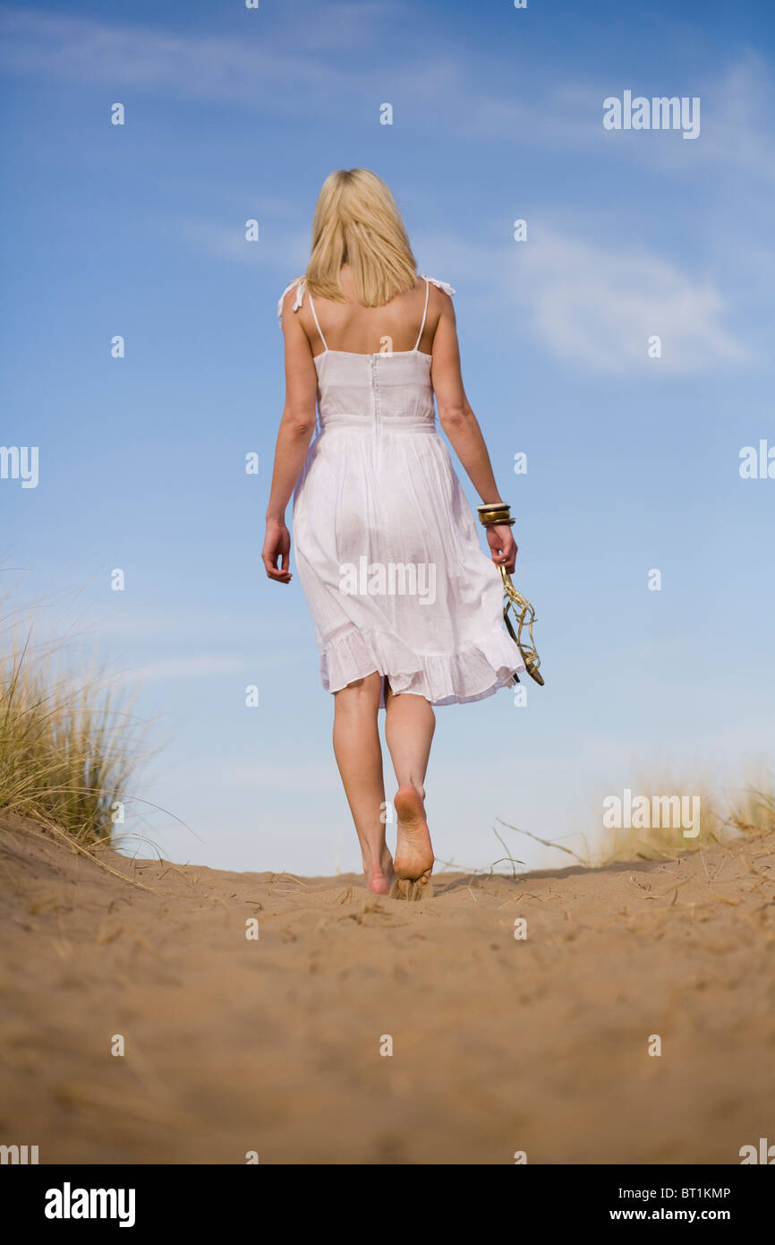 Una giovane donna bionda si allontana a piedi nudi nelle dune di sabbia che porta i suoi sandali in una giornata calda Foto stock Alamy