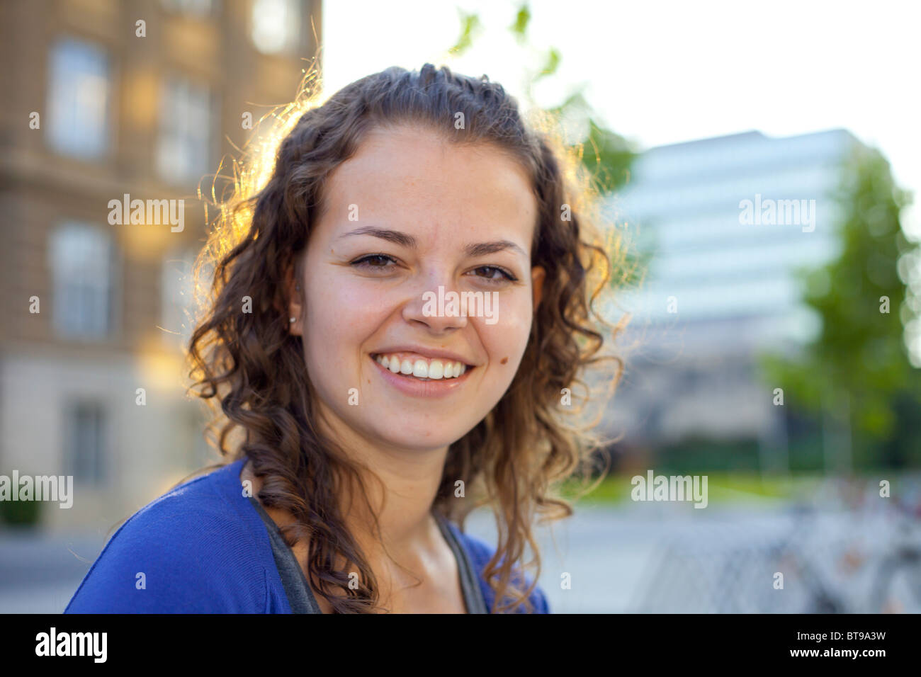 La giovane donna con i capelli castani ricci sorride calorosamente all'aperto, sotto la luce del sole soffusa, in piedi di fronte a uno sfondo urbano. Foto Stock