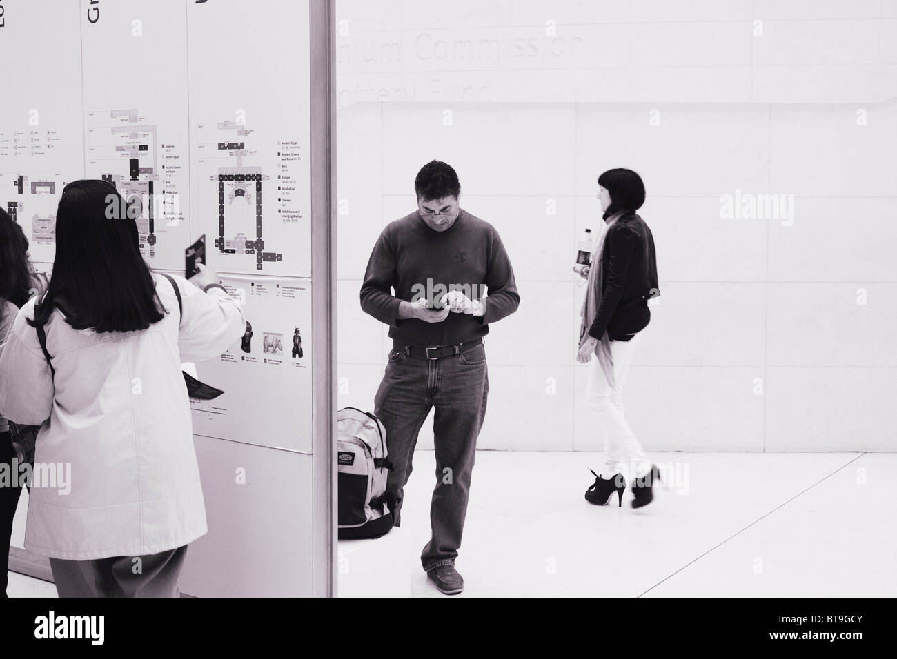 Un uomo guarda al suo telefono mentre alcune persone guardano il piano guida e un'altra donna da passeggiate nel British Museum di Atrium. Foto Stock
