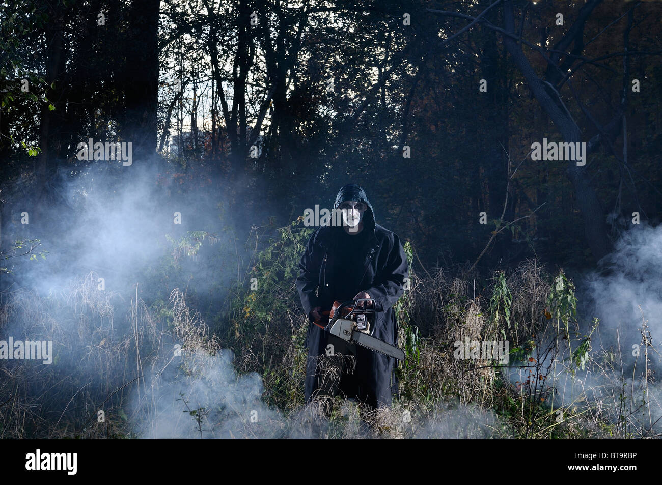 Clamorosa male pallido man in black coat con una sega a nastro in un bosco nebbioso nella caduta di halloween Foto Stock