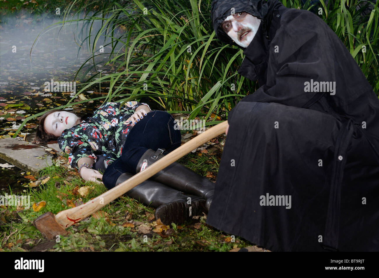 Micidiali uomo malvagio nel mantello nero e bianco di fronte con ax accovacciato su un dead girls corpo mediante un laghetto Foto Stock
