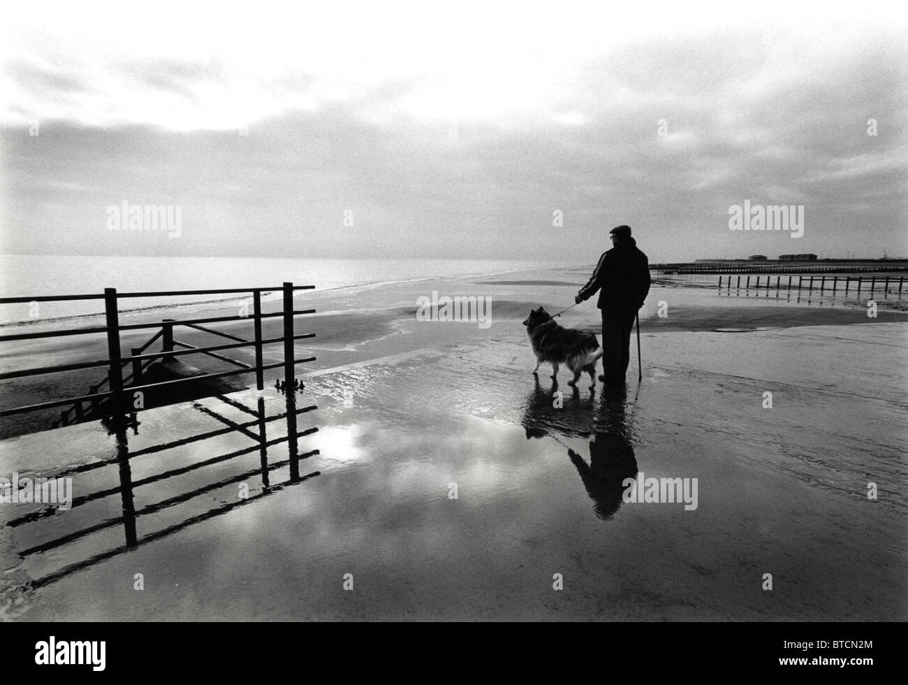 Un uomo anziano con il suo cane su una spiaggia deserta con un molo in background Foto Stock