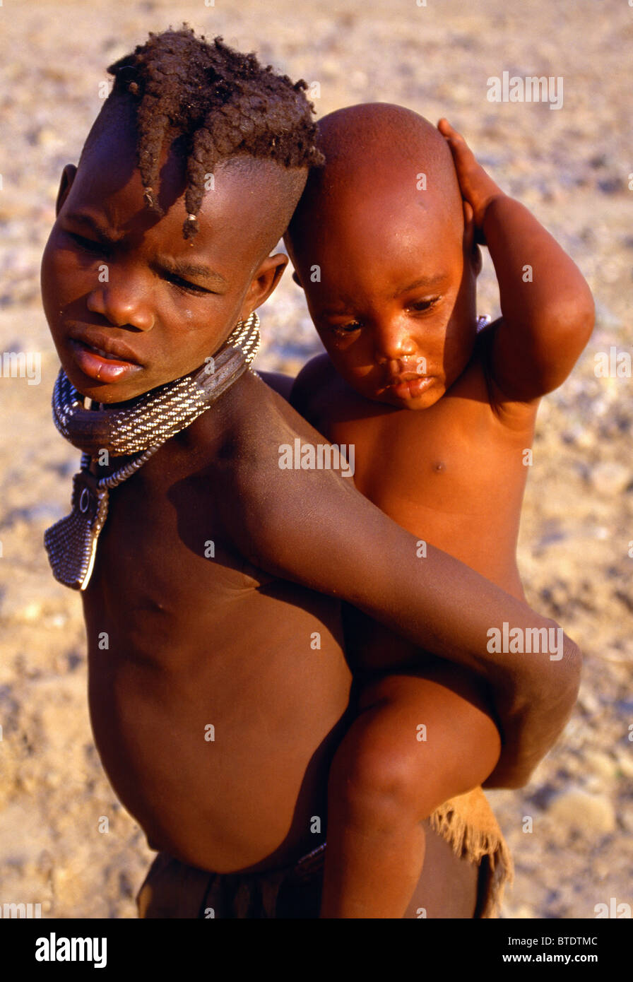 Giovane ragazzo Himba con intrecciato di acconciatura portando un giovane gemelli sulla sua schiena Foto Stock