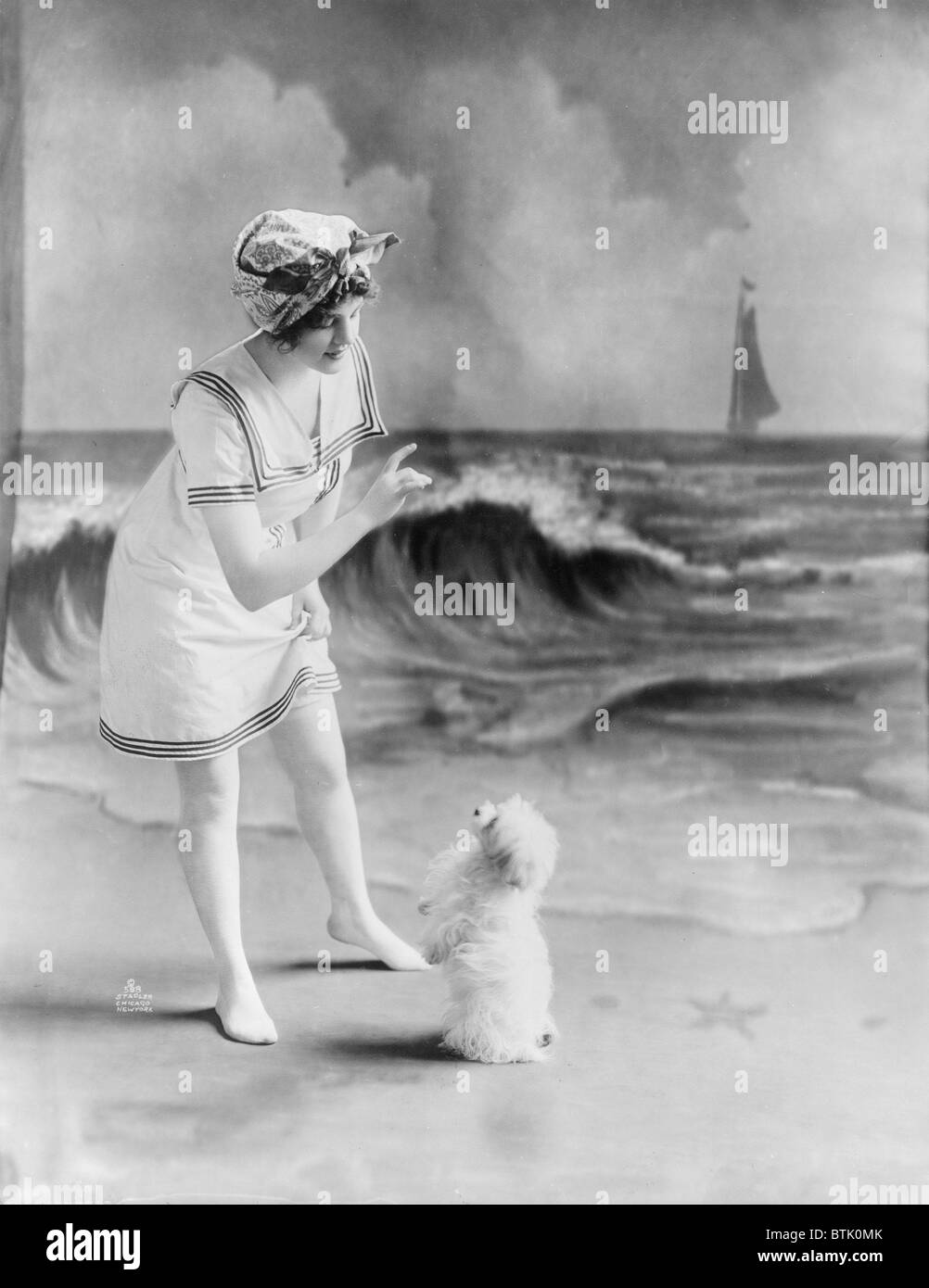 Bellezza balneare, giovane donna in costume da bagno con un cane di piccola taglia di fronte studio sullo sfondo della spiaggia, fotografia, 1913. Foto Stock