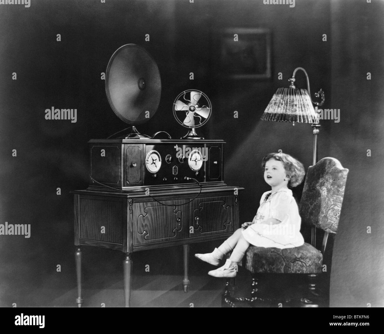 Poco ragazza seduta su una sedia accanto ad una alimentazione elettrica  radio, con un ventilatore elettrico e luce elettrica. 1926 Foto stock -  Alamy