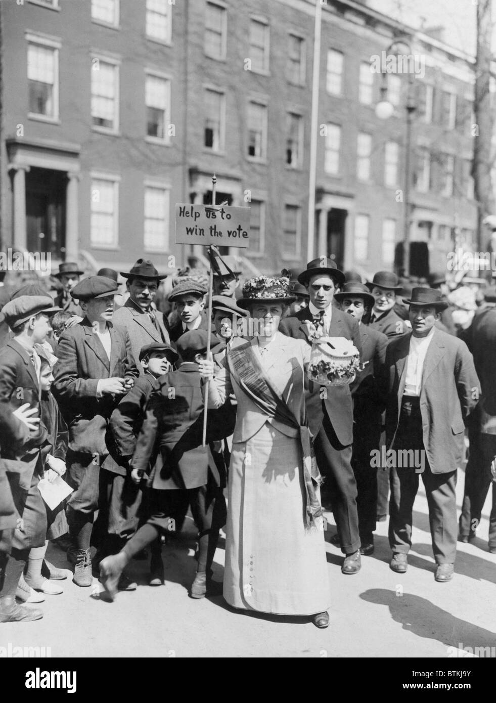 Le donne identificate come la sig.ra Suffern, è circondato da una folla di uomini e ragazzi, mentre lei detiene un banner in donne suffragist parade "Aiutaci a vincere la votazione." 1914. Foto Stock
