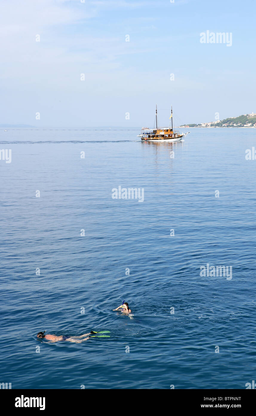Persone fare snorkelling nelle acque limpide del mare Adriatico mentre una barca pases da vicino alla città di Omis Croazia Foto Stock