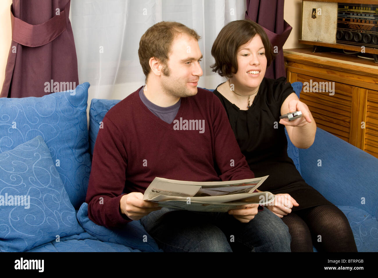 Un giovane si siede su un divano blu nel loro salotto con un quotidiano e un telecomando TV. La donna è rivolta a qualcosa sulla TV. Foto Stock