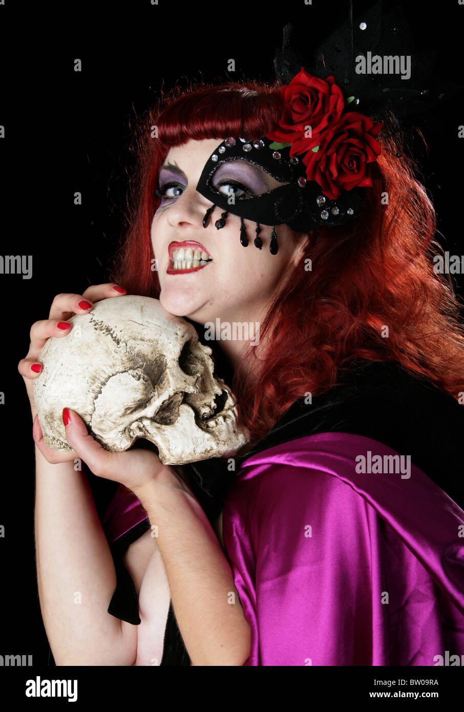 Rosso giovane intitolata donna che indossa un capo di colore viola e maschera Sequined su di un occhio e una Rosa Rossa e tenendo un teschio umano Foto Stock