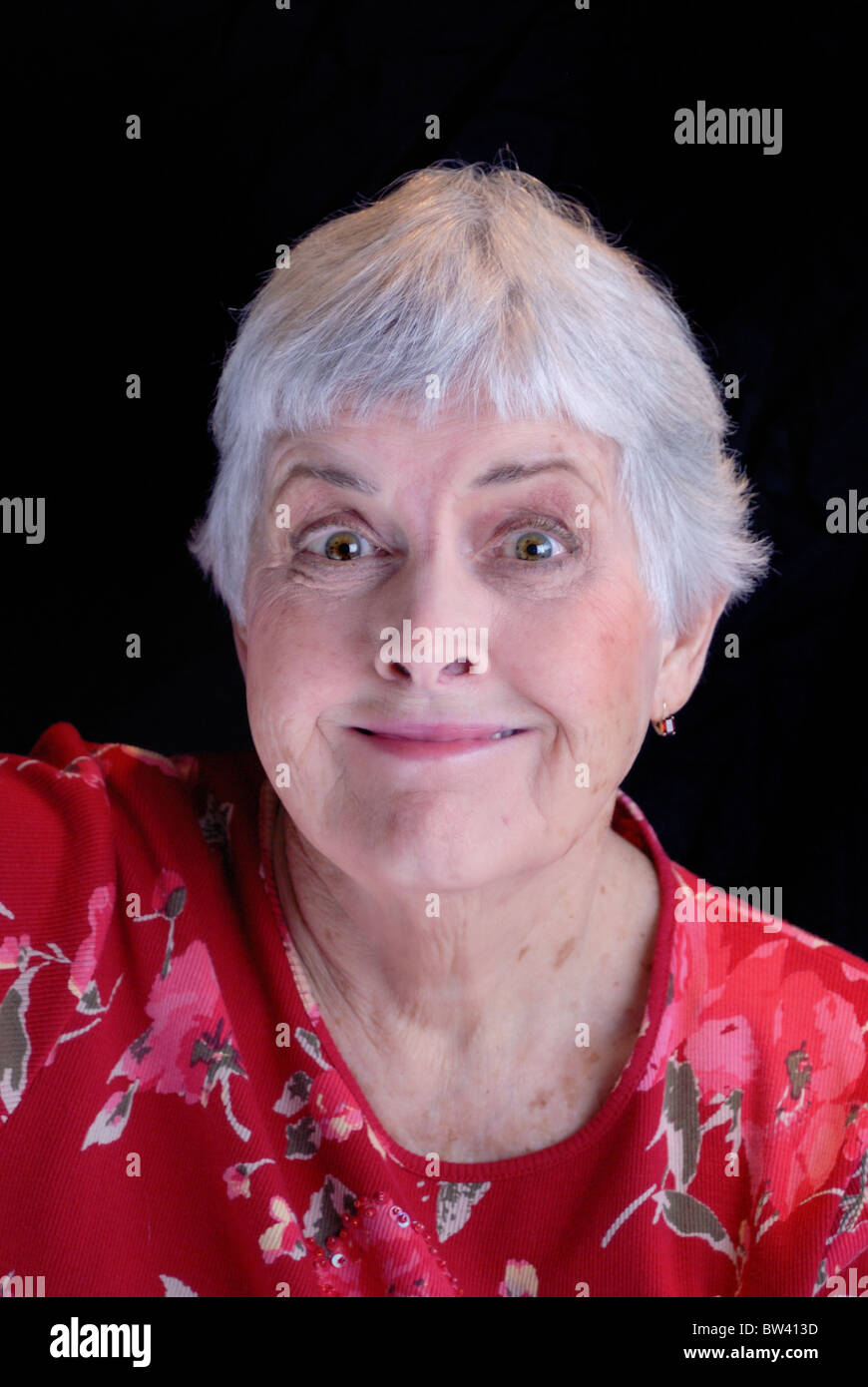 Un vibrante senior citizen donna con i capelli bianchi che indossa un fiore rosso shirt rende una buffa faccia con un pippo sorriso e gli occhi grandi. Foto Stock