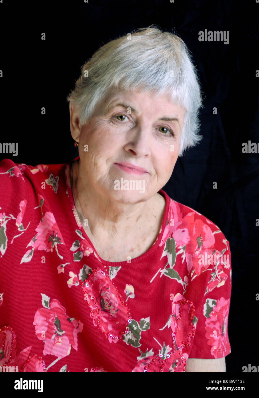 Un vibrante senior citizen donna witih capelli bianchi che indossa un fiore rosso camicia e sorridente. Foto Stock