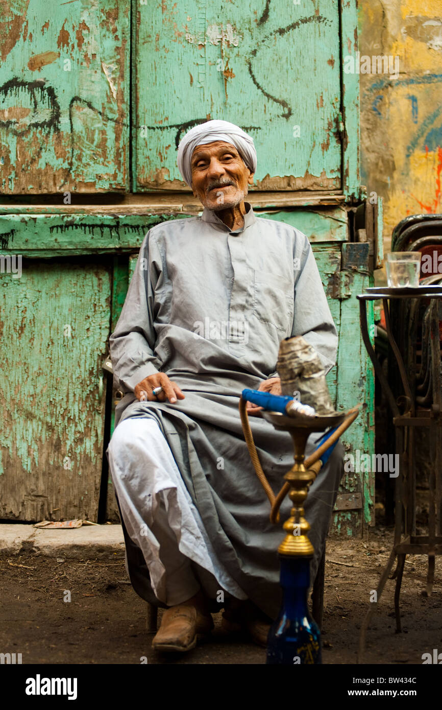 Un amichevole vecchio uomo egiziano siede a un street cafe fumare narghilè in Cairo islamico, Egitto Foto Stock