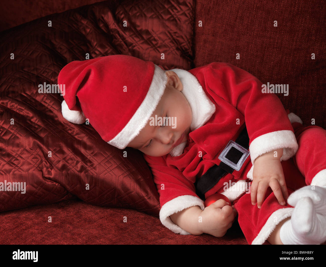 Licenza disponibile all'indirizzo MaximImages.com - bambino di sei mesi in costume di Babbo Natale che dorme su un divano con un bauble di Natale in mano Foto Stock