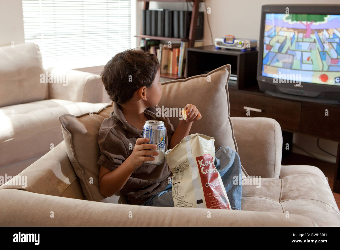 Quattro-anno-vecchio ragazzo Mexican-American mangia patatine, sips soft drink e guarda la TV in salotto nella sua casa di Austin Foto Stock