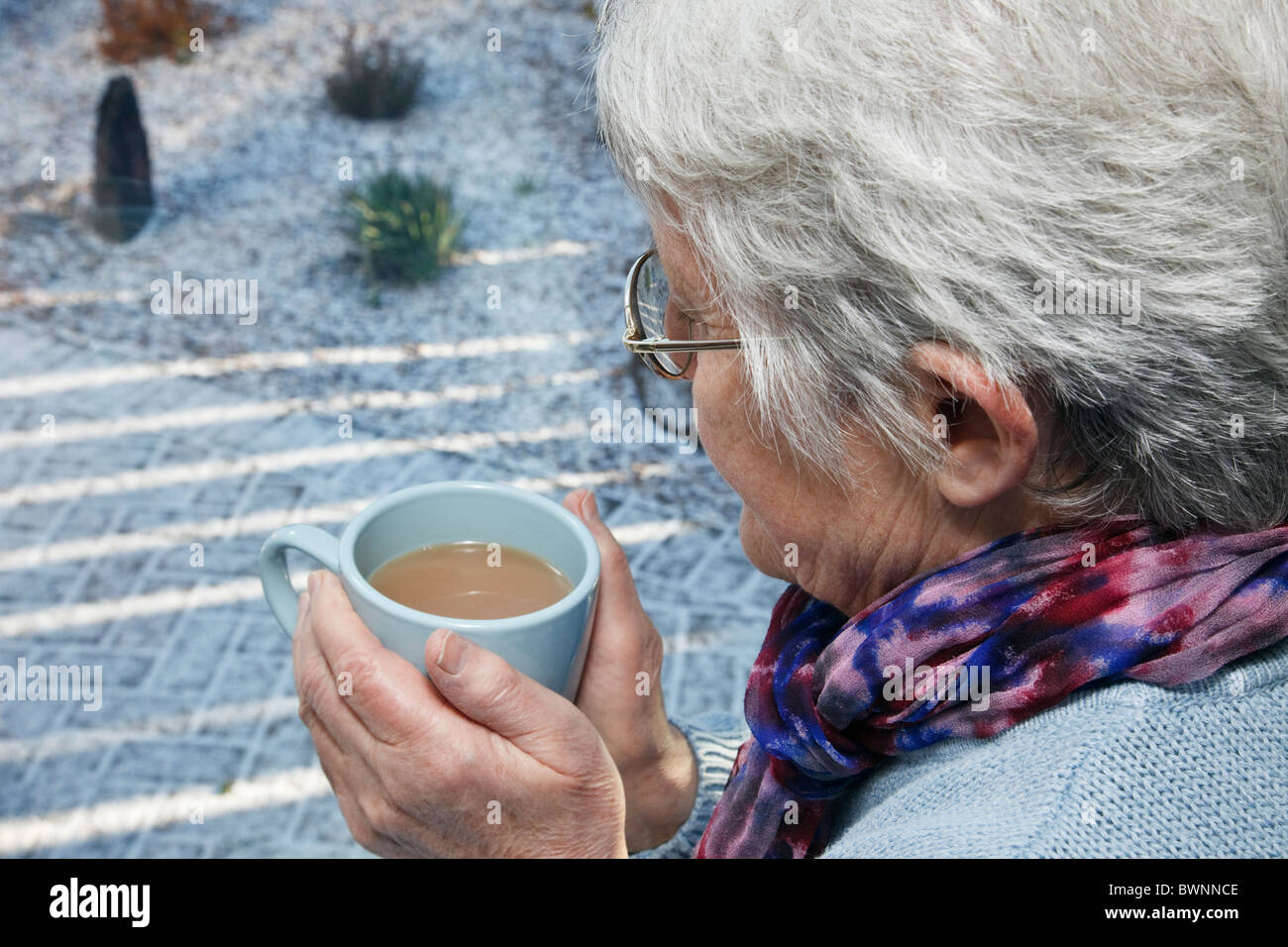 Hygge quotidiana scena con alti donna sola indossando una sciarpa e tenendo una bevanda calda guardando fuori della finestra in un freddo giorno di neve in inverno. Regno Unito Foto Stock